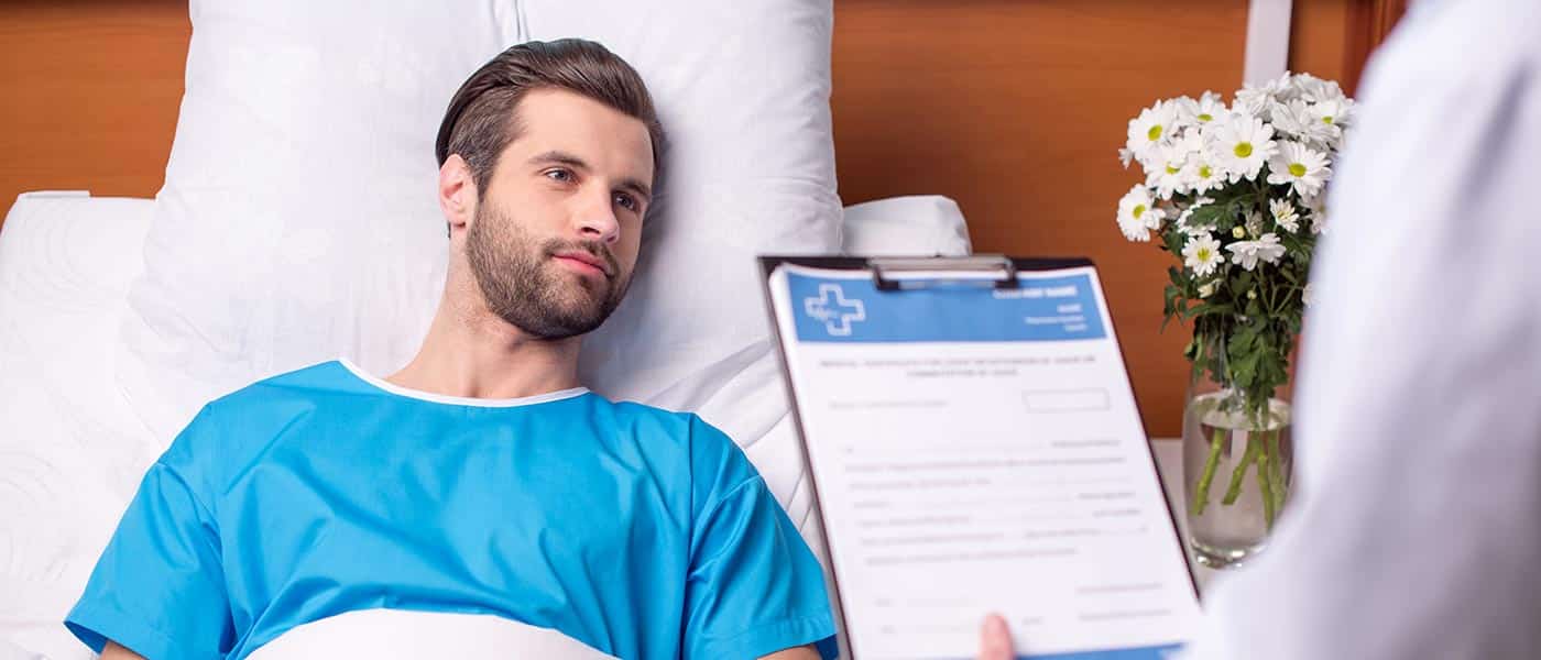 Patient in a blue hospital gown lying in bed next to flowers while doctor holds clipboard.