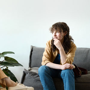 Person sitting on gray sofa wearing a tan jacket and jeans, looking thoughtful.