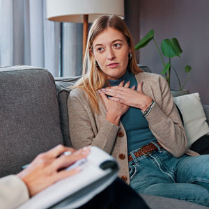 Person in a beige cardigan sitting on a couch with hands on chest, conversing with unseen person holding notebook.