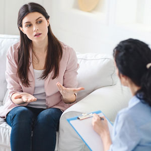 Person sitting on a couch wearing a pink cardigan, gesturing while talking to another person taking notes.