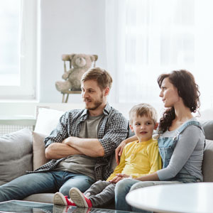 Family sitting on couch, man in checkered shirt, woman in gray top, child in yellow shirt.